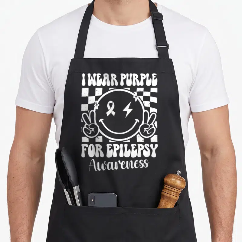 Epilepsy face apron - White smiley face with ribbon lightning bolt and peace signs on checkerboard with awareness text — shown on a black cotton canvas apron against a white background