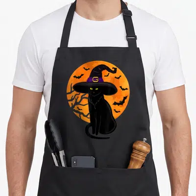 Pumpkin black cat apron - Cartoon black cat wearing purple witch hat sitting before orange moon with bats and tree — shown on a black cotton canvas apron against a white background