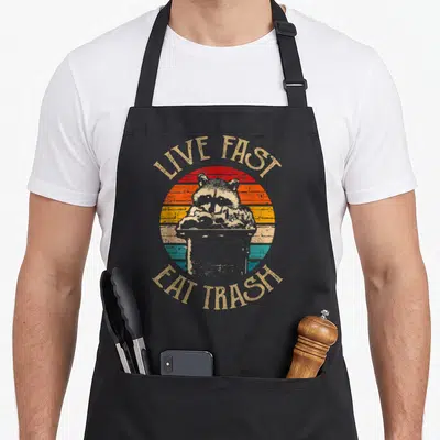 Forest cycling apron - Raccoon in trash can with arched LIVE FAST EAT TRASH text over retro sun stripes — shown on a black cotton canvas apron against a white background