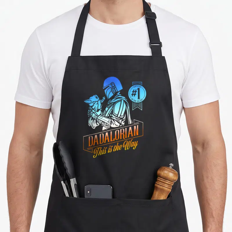 Funny galaxy father day apron - Cartoon Mandalorian holding Grogu with Dadalorian text and certified number one seal — shown on a black cotton canvas apron against a white background