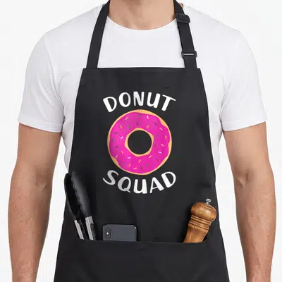 Squad donut apron - Pink frosted donut with sprinkles between white DONUT and SQUAD text — shown on a black cotton canvas apron against a white background