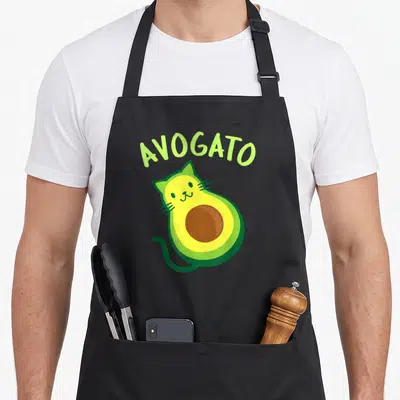 Cinco avocado apron - Arched AVOGATO text above a cartoon avocado cat in lime green, yellow, and brown — shown on a black cotton canvas apron against a white background