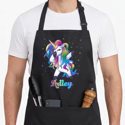 Adley rainbow apron - Cartoon unicorn dabbing with rainbow mane and Adley text below surrounded by stars — shown on a black cotton canvas apron against a white background