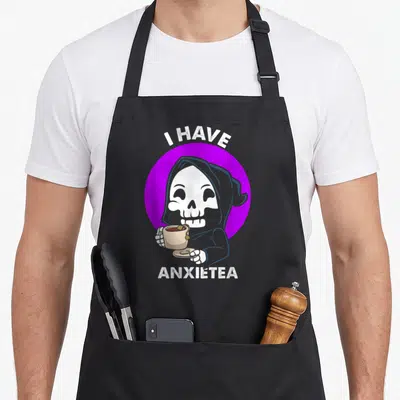 Goth tea apron - Cartoon grim reaper in purple circle holding beige teacup with I Have Anxietea text — shown on a black cotton canvas apron against a white background