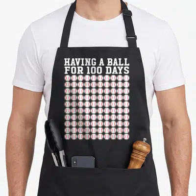 Player teacher apron - Having A Ball For 100 Days text above 100 baseballs with red stitching — shown on a black cotton canvas apron against a white background