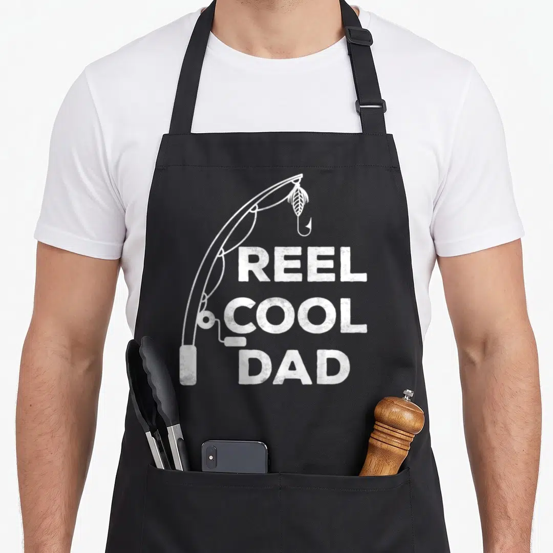 Father fisherman apron - White distressed fishing rod curving over stacked Reel Cool Dad text — shown on a black cotton canvas apron against a white background