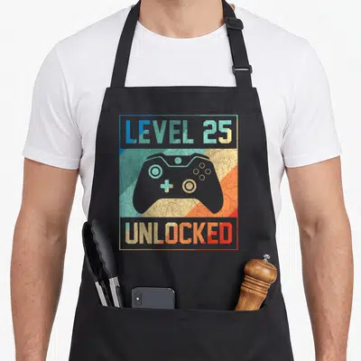 25th birthday apron - Retro grey game controller with LEVEL 25 UNLOCKED text over multicolored diagonal stripes — shown on a black cotton canvas apron against a white background