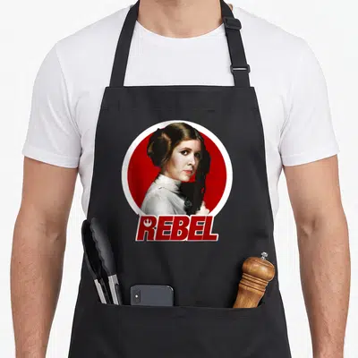 Original princess apron - Princess Leia portrait in red circle with REBEL text below — shown on a black cotton canvas apron against a white background