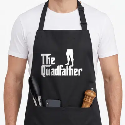 Quadfather leg apron - White The Quadfather text with muscular leg silhouette graphic — shown on a black cotton canvas apron against a white background