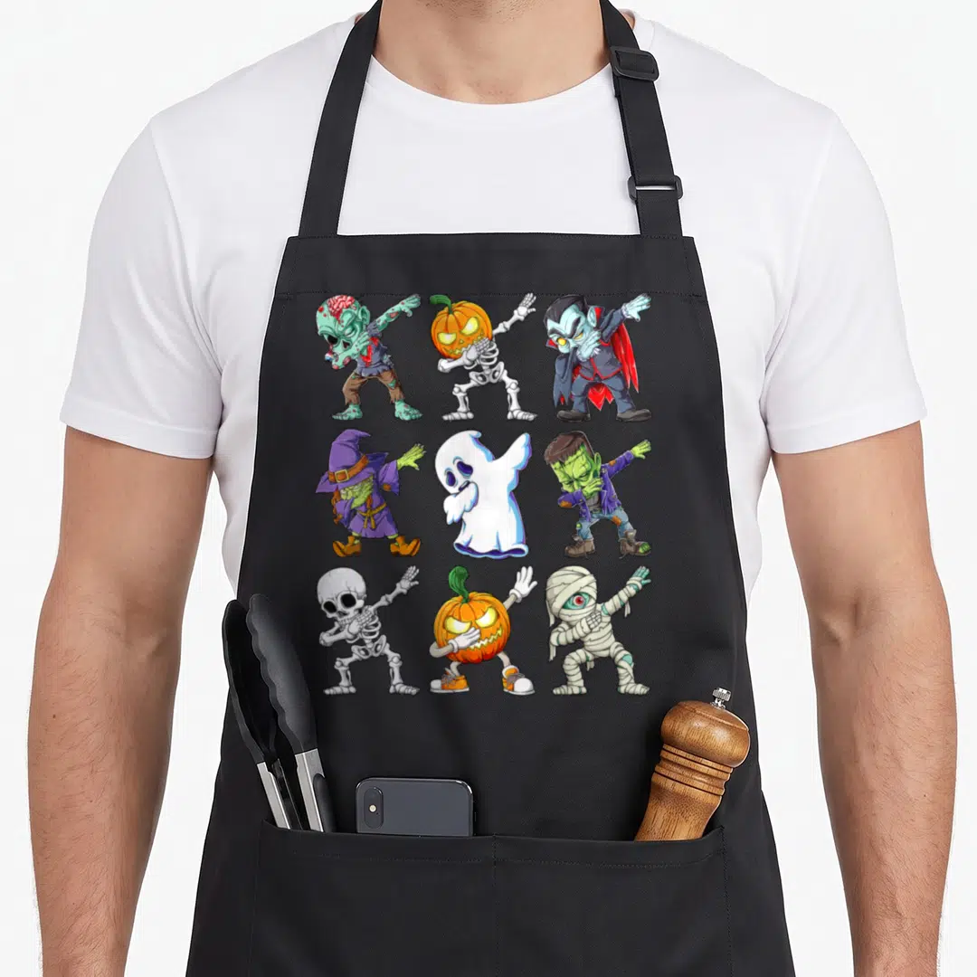 Dabbing halloween kitchen aprons - Nine cartoon monsters including zombie, skeleton, pumpkin, vampire, witch, ghost — shown on a black cotton canvas apron against a white background