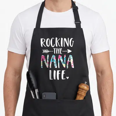 Rocking grandma apron - ROCKING THE NANA LIFE. text with floral NANA and arrow — shown on a black cotton canvas apron against a white background