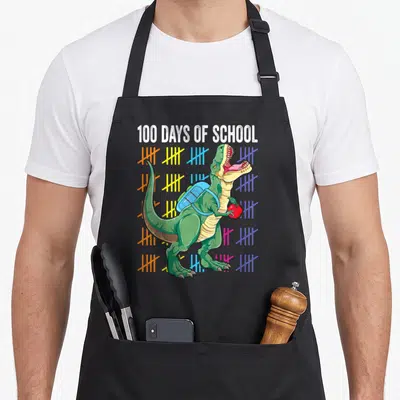 100th t-rex apron - Green T-Rex with blue backpack and red apple surrounded by colorful tally marks under white text — shown on a black cotton canvas apron against a white background