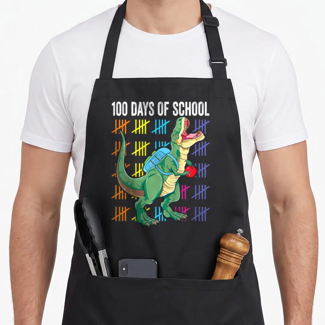 100th t-rex apron - Green T-Rex with blue backpack and red apple surrounded by colorful tally marks under white text — shown on a black cotton canvas apron against a white background