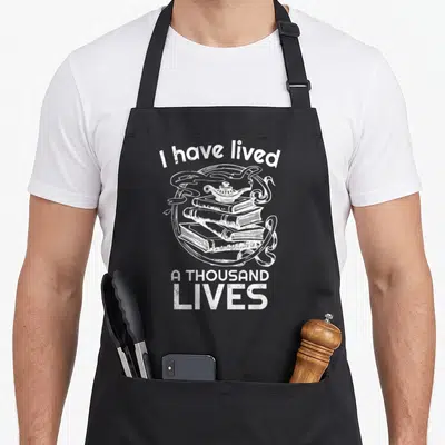 Lived bestseller apron - White line art of stacked books with magic lamp and arched text I have lived A THOUSAND LIVES — shown on a black cotton canvas apron against a white background