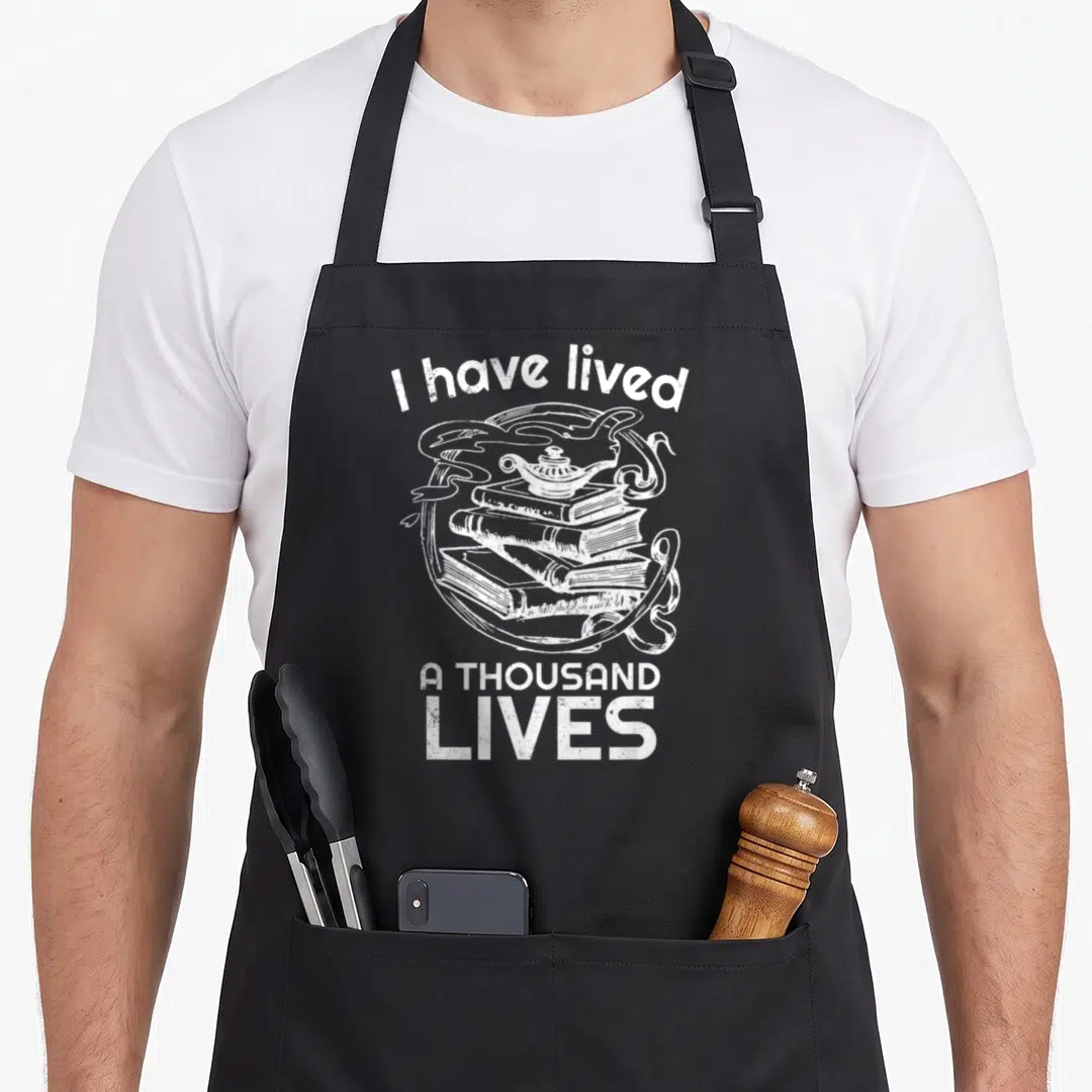 Lived bestseller apron - White line art of stacked books with magic lamp and arched text I have lived A THOUSAND LIVES — shown on a black cotton canvas apron against a white background