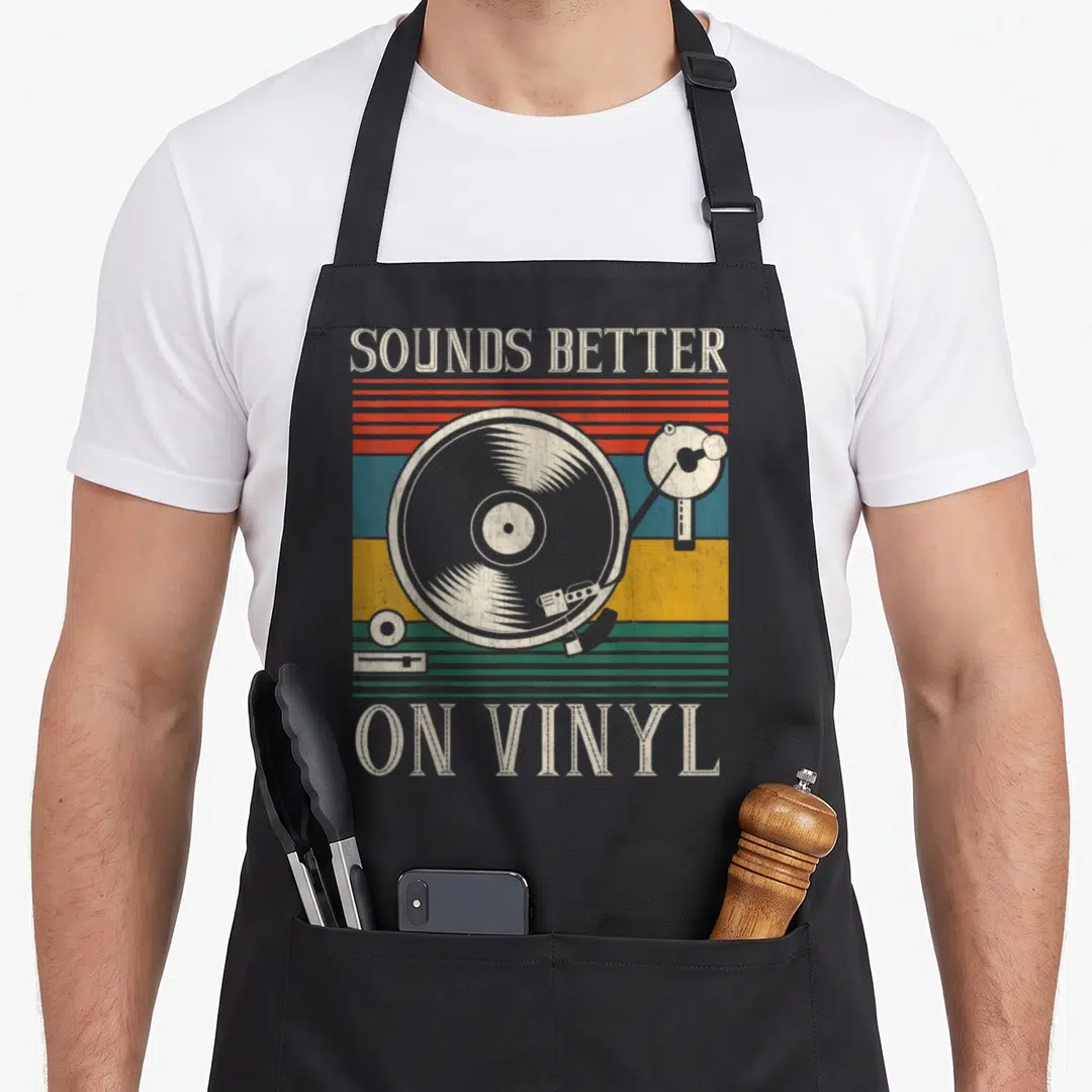 Sound better vinyl apron - Retro record player illustration with striped background and SOUNDS BETTER ON VINYL text — shown on a black cotton canvas apron against a white background