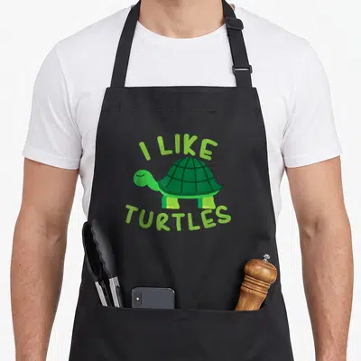 Turtle animal apron - Green cartoon turtle centered between arched I LIKE TURTLES text — shown on a black cotton canvas apron against a white background