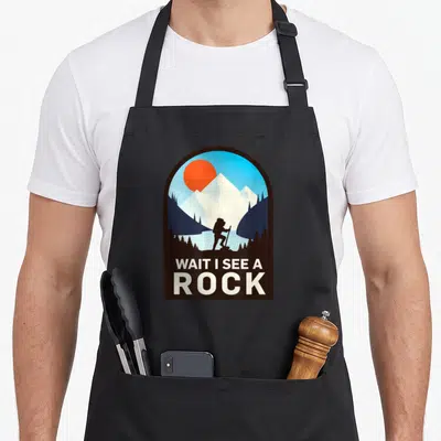 Wait geologist lover apron - Retro hiker silhouette under brown arch with text WAIT I SEE A ROCK in white — shown on a black cotton canvas apron against a white background