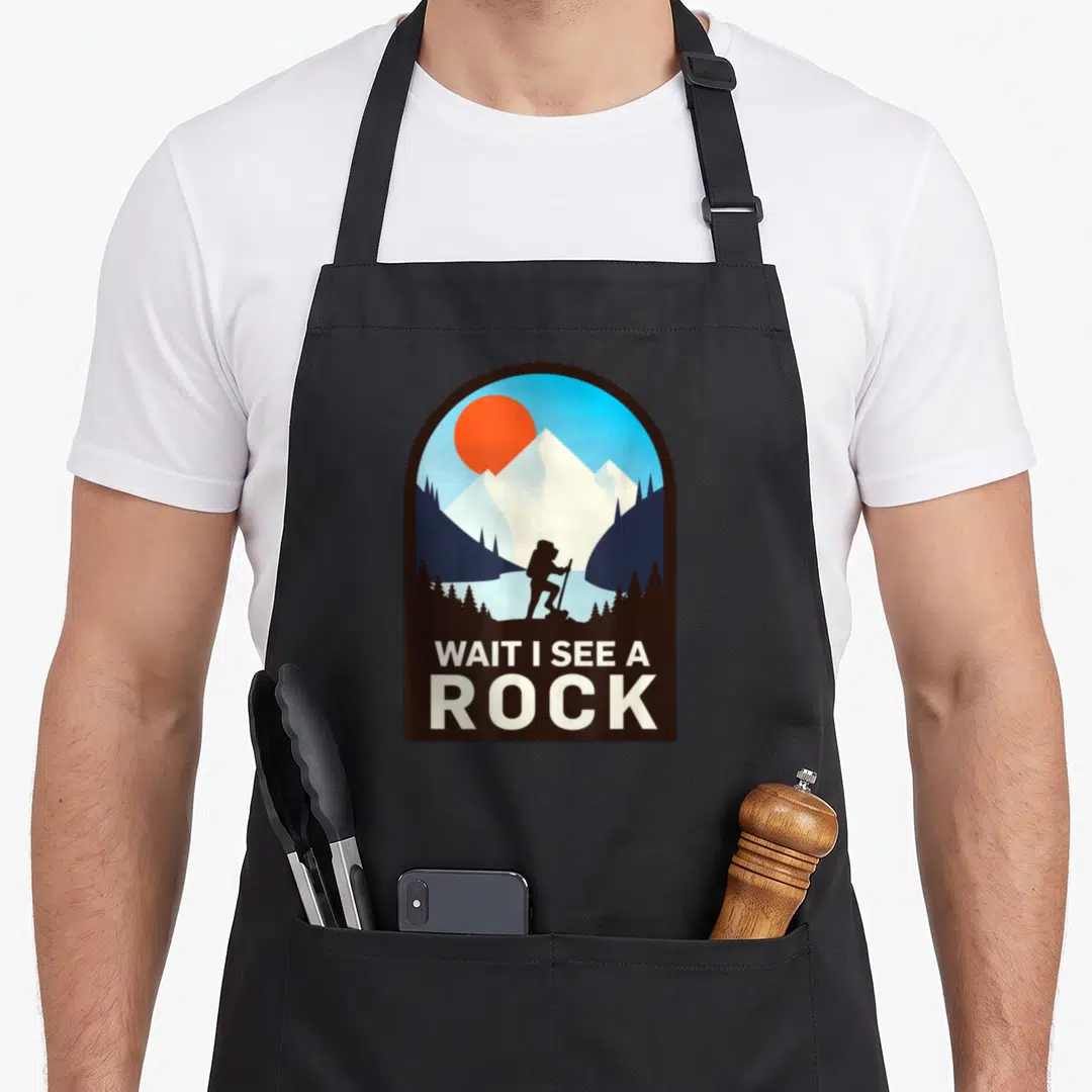 Wait geologist lover apron - Retro hiker silhouette under brown arch with text WAIT I SEE A ROCK in white — shown on a black cotton canvas apron against a white background