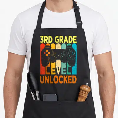 3rd video game apron - Retro controller over yellow orange red blue grey stripes with 3rd Grade Level Unlocked text — shown on a black cotton canvas apron against a white background