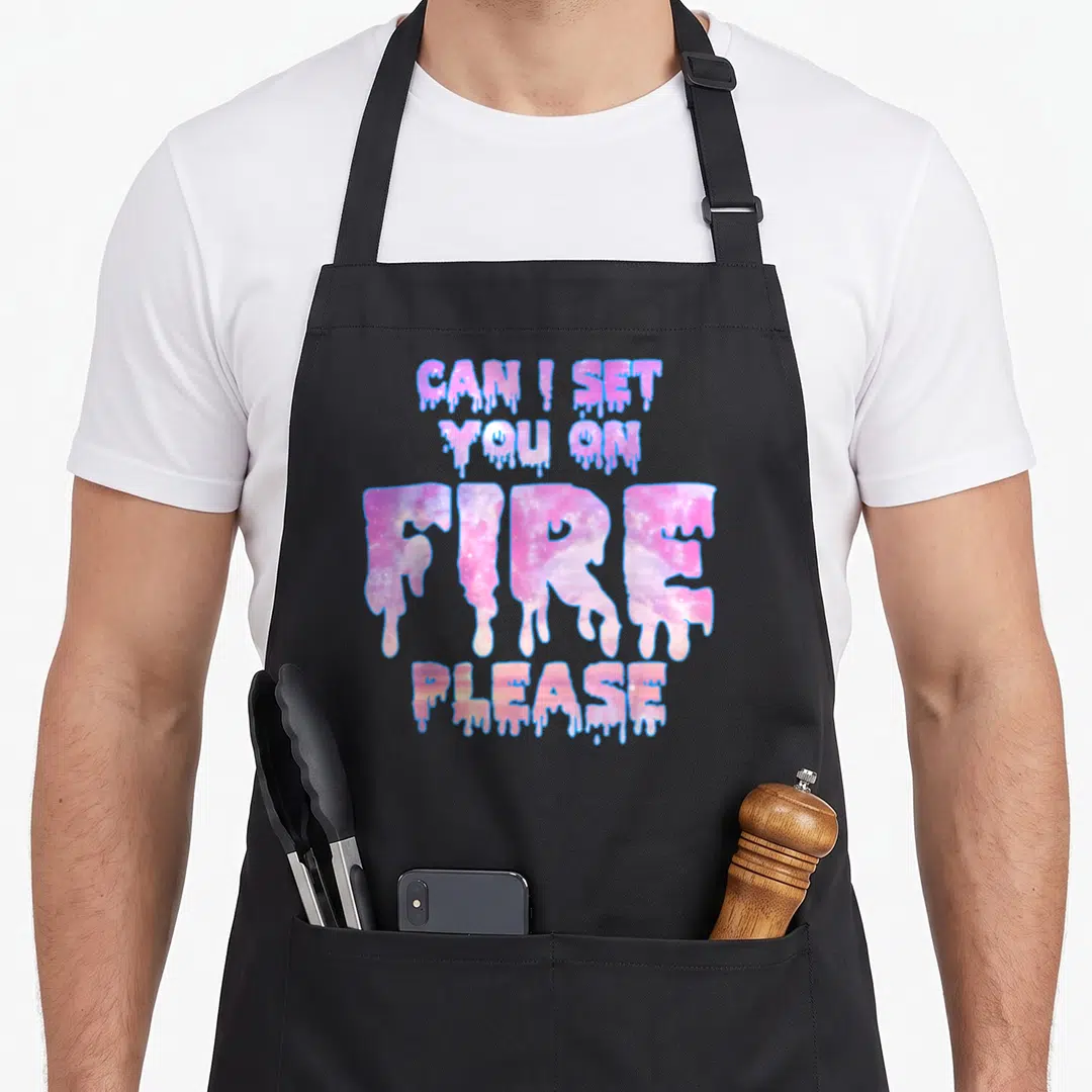 Pastel snarky aprons - Dripping galaxy text reading CAN I SET YOU ON FIRE PLEASE in pink and purple — shown on a black cotton canvas apron against a white background