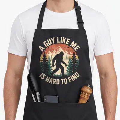 Cute hard bigfoot apron - Retro bigfoot silhouette in orange sunset with pine trees and arched text — shown on a black cotton canvas apron against a white background