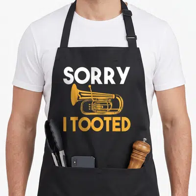 Tooted i love tuba apron - Orange cartoon tuba between white SORRY and orange I TOOTED text — shown on a black cotton canvas apron against a white background