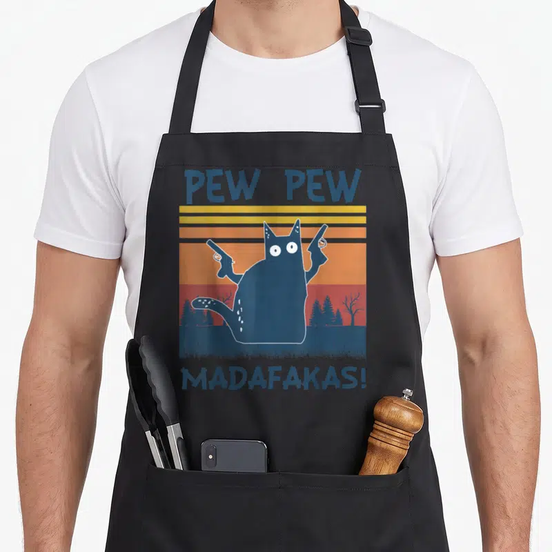 Pew madafakas cat apron - Cartoon cat holding two guns against striped sunset with PEW PEW MADAFAKAS text — shown on a black cotton canvas apron against a white background