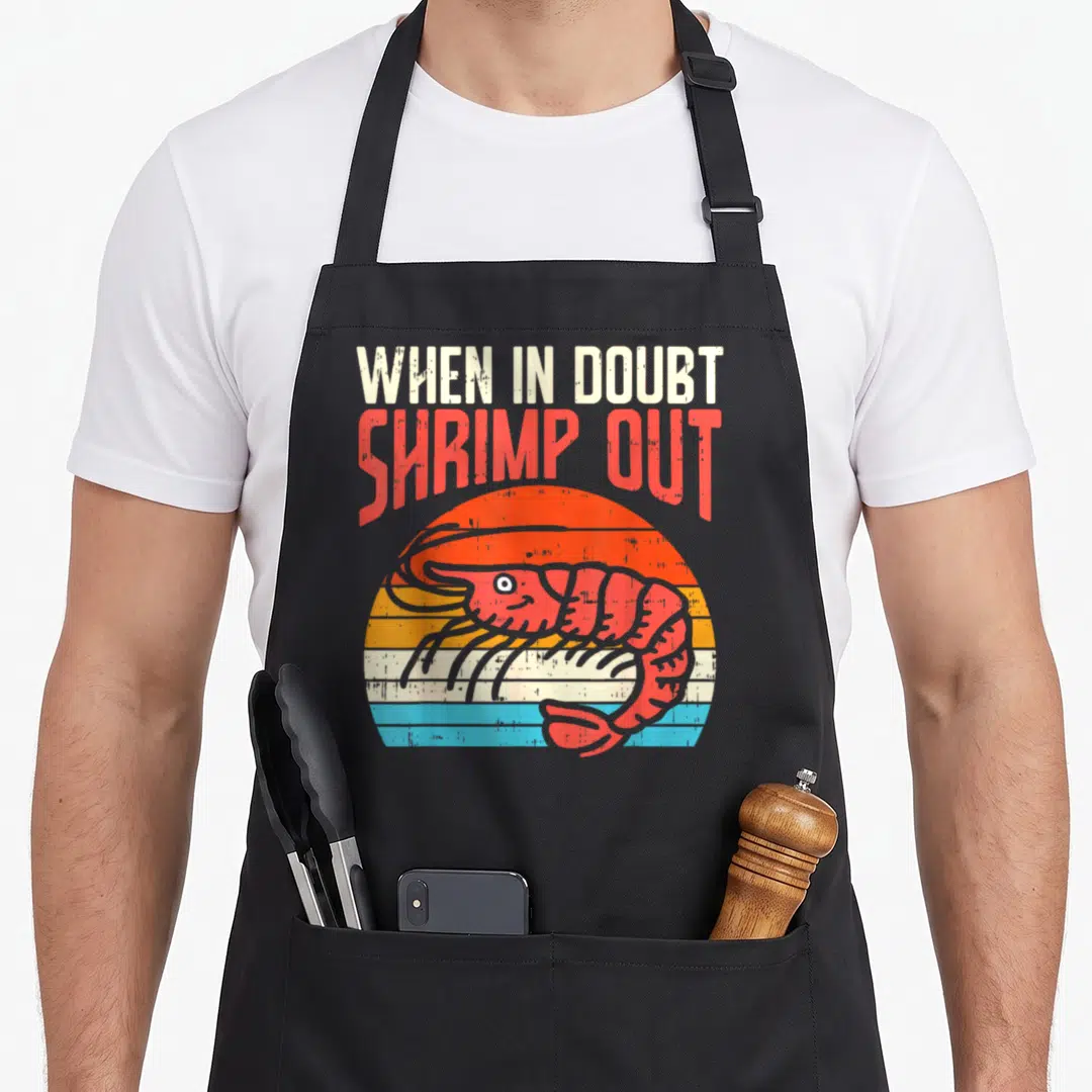 Doubt boredkoalas apron - Red shrimp centered on a striped orange and blue sun with stacked text reading When In Doubt — shown on a black cotton canvas apron against a white background