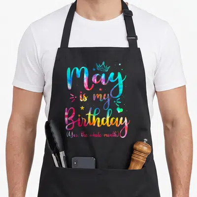 May tie-dye apron - Tie-dye text reading May is my Birthday with crown and stars in pink blue yellow and green — shown on a black cotton canvas apron against a white background