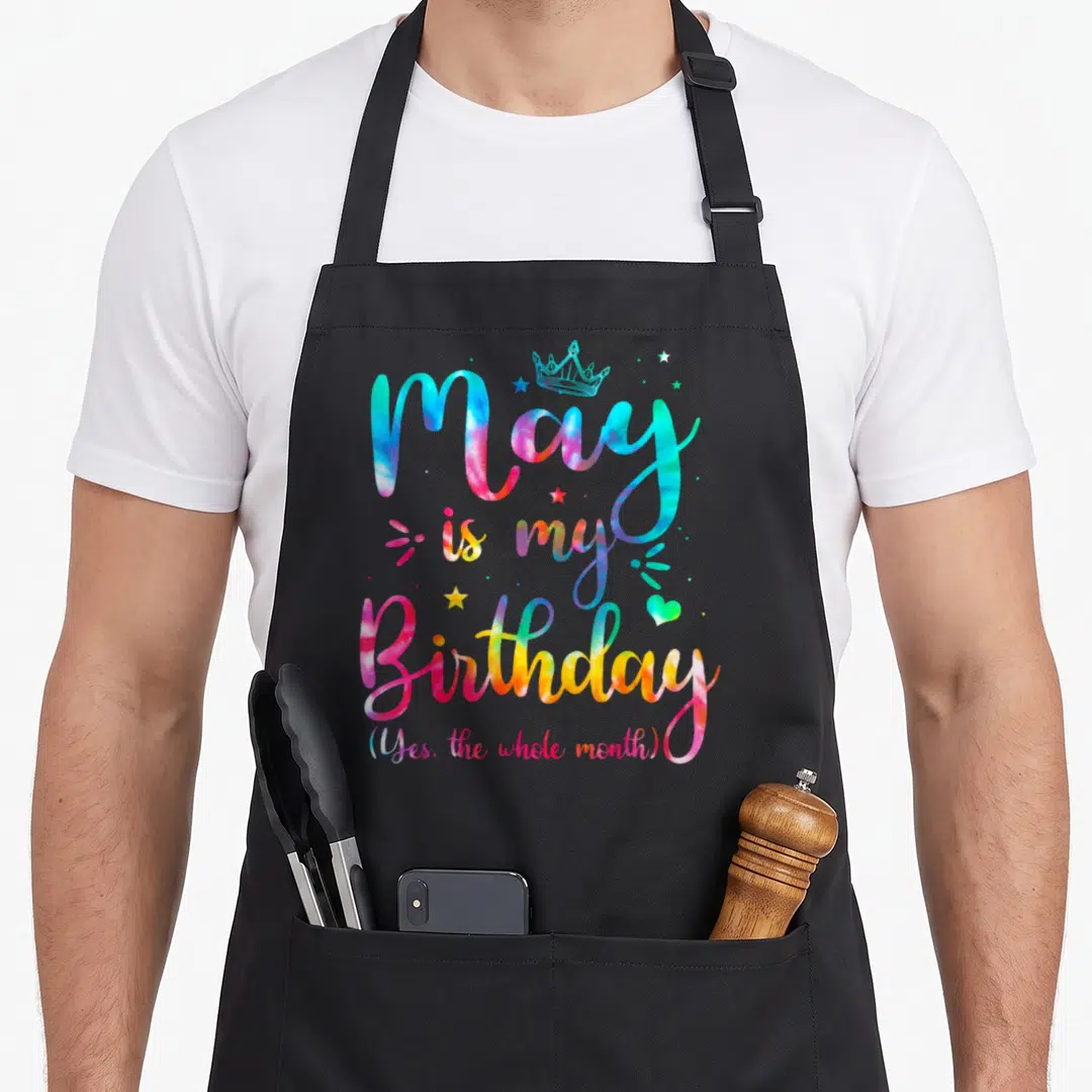 May tie-dye apron - Tie-dye text reading May is my Birthday with crown and stars in pink blue yellow and green — shown on a black cotton canvas apron against a white background