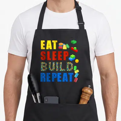 Sleep grill master apron - EAT SLEEP BUILD REPEAT text formed by yellow red blue and green plastic building blocks — shown on a black cotton canvas apron against a white background