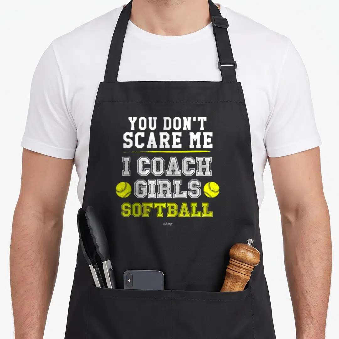 Softball panic attack apron - Stacked white and yellow text I COACH GIRLS SOFTBALL with two yellow softballs flanking — shown on a black cotton canvas apron against a white background