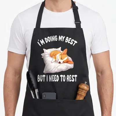 Sleeping corgi aprons - Orange and white corgi sleeping on white pillow with text I'M DOING MY BEST, BUT I NEED TO REST — shown on a black cotton canvas apron against a white background