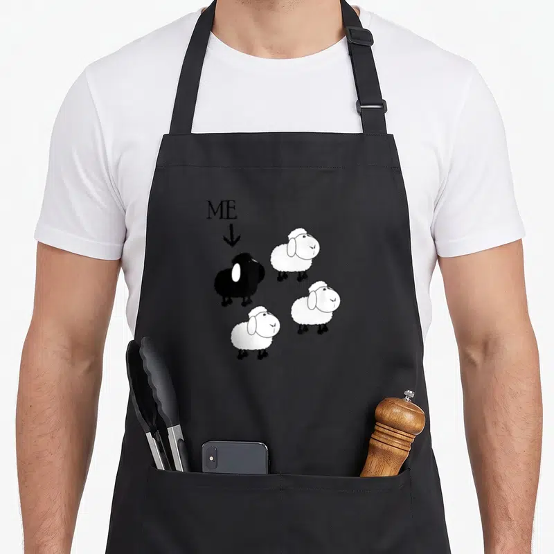 Funny family sheep apron - ME label pointing to black sheep among three white sheep cartoon — shown on a black cotton canvas apron against a white background