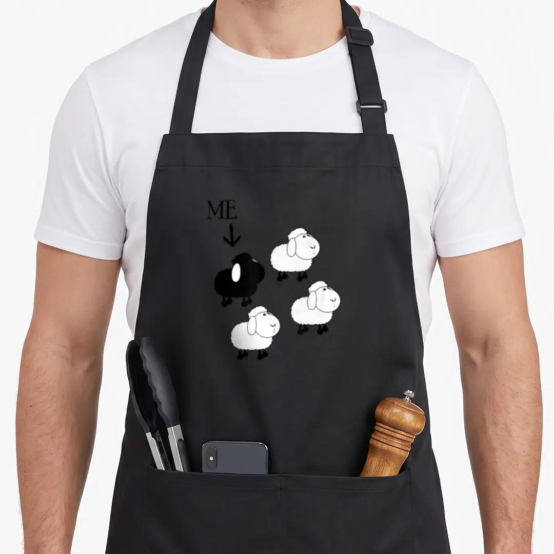 Funny family sheep apron - ME label pointing to black sheep among three white sheep cartoon — shown on a black cotton canvas apron against a white background