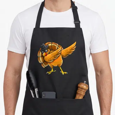 Dabbing thanksgiving day aprons - Cartoon turkey in black hat and sunglasses doing a dab pose in orange and yellow — shown on a black cotton canvas apron against a white background
