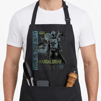 Way ivorian apron - The Mandalorian standing with Grogu in a pod and TIE fighters under distressed blue and green text — shown on a black cotton canvas apron against a white background