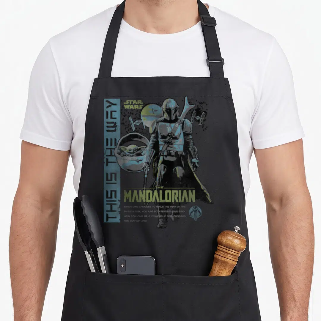 Way ivorian apron - The Mandalorian standing with Grogu in a pod and TIE fighters under distressed blue and green text — shown on a black cotton canvas apron against a white background