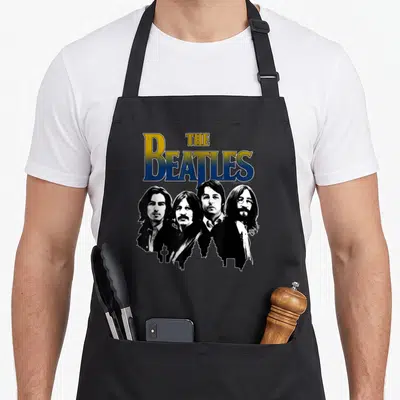 Funny skyline apron - The Beatles text above four portraits and black city skyline silhouette — shown on a black cotton canvas apron against a white background