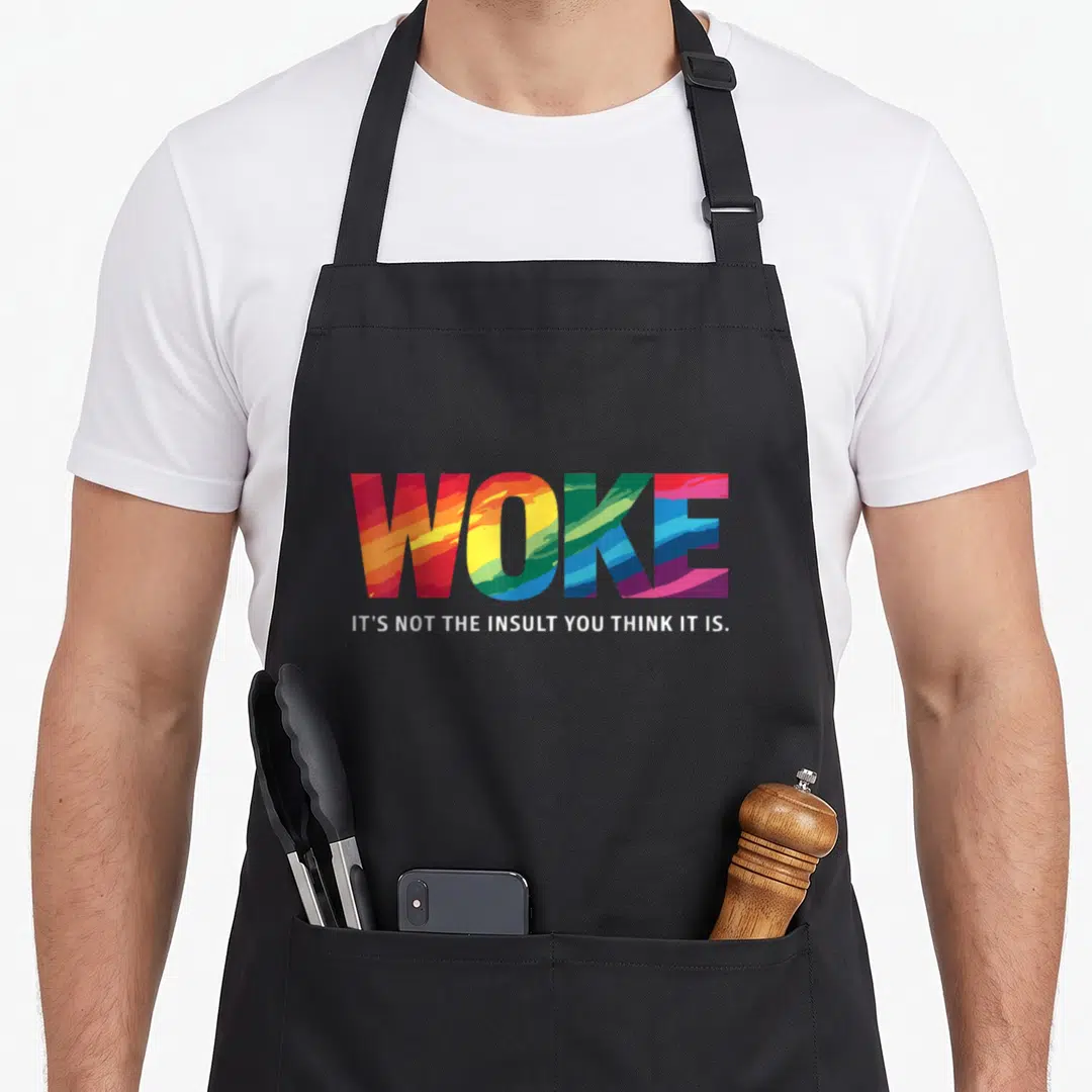 Woke novelty apron - Rainbow brushed WOKE text above white sentence IT'S NOT THE INSULT YOU THINK IT IS — shown on a black cotton canvas apron against a white background