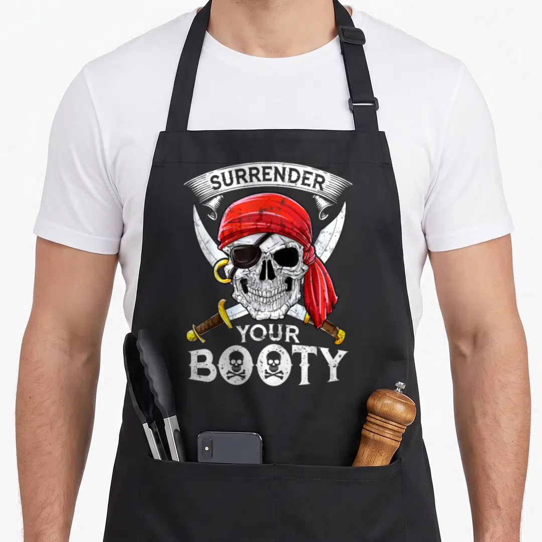 Surrender skull apron - Pirate skull with red bandana over crossed swords and arched text — shown on a black cotton canvas apron against a white background