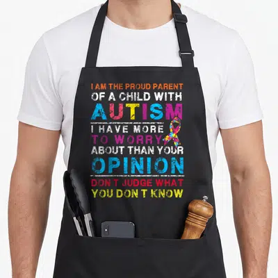 Proud judge dad apron - Multi-colored text reading I am the proud parent of a child with autism next to a puzzle ribbon — shown on a black cotton canvas apron against a white background