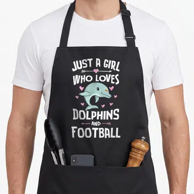 Dolphin football apron - Cartoon dolphin surrounded by pink hearts with text Just A Girl Who Loves Dolphins And Football — shown on a black cotton canvas apron against a white background