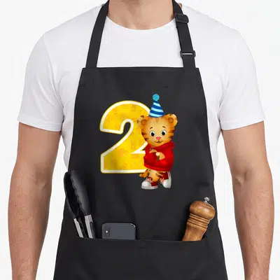 Daniel 2nd apron - Cartoon tiger in red sweater next to yellow number 2 with blue hat — shown on a black cotton canvas apron against a white background