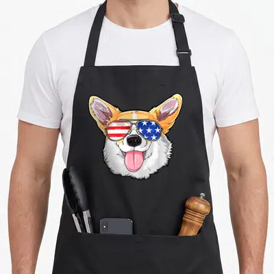 Corgi 4th of july apron - Cartoon corgi face with red white and blue star stripe sunglasses — shown on a black cotton canvas apron against a white background
