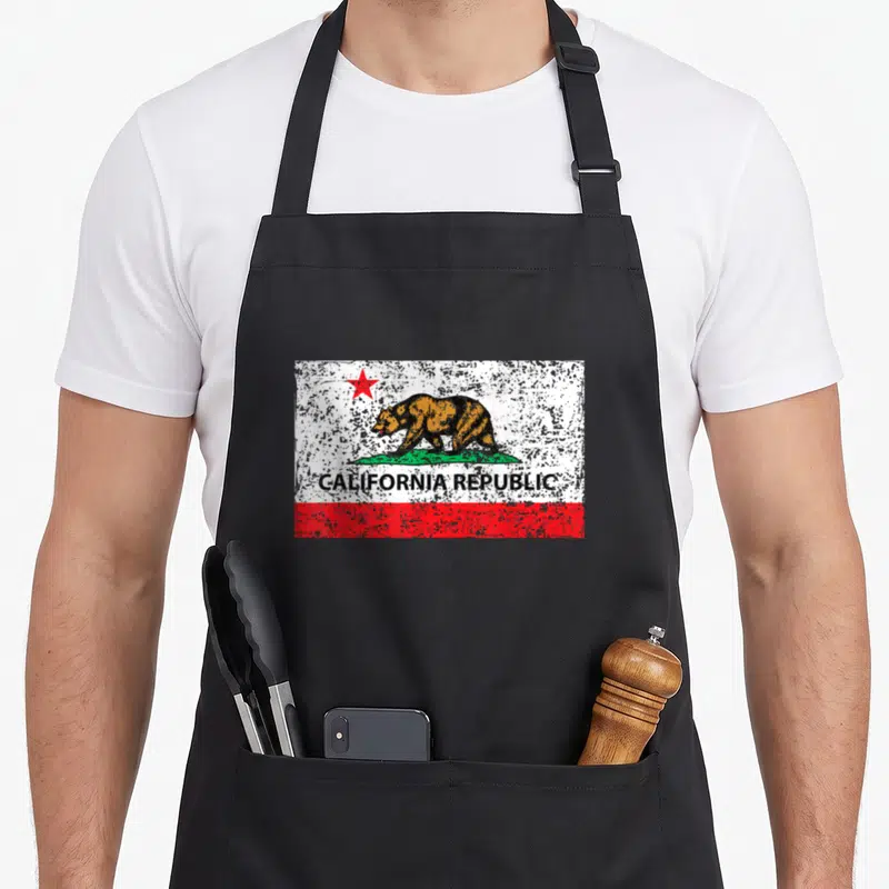 Republic california apron - Distressed California flag with brown bear, red star, green grass, and black text on white — shown on a black cotton canvas apron against a white background