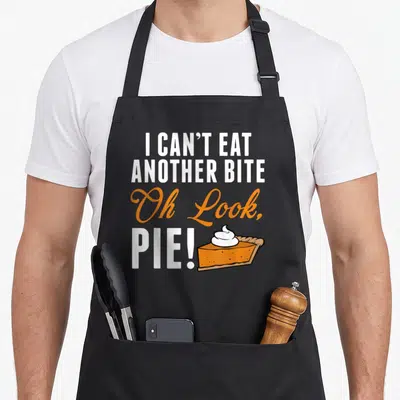 Eat pie apron - Stacked text I CAN'T EAT ANOTHER BITE Oh Look PIE next to an orange pie slice with white cream — shown on a black cotton canvas apron against a white background