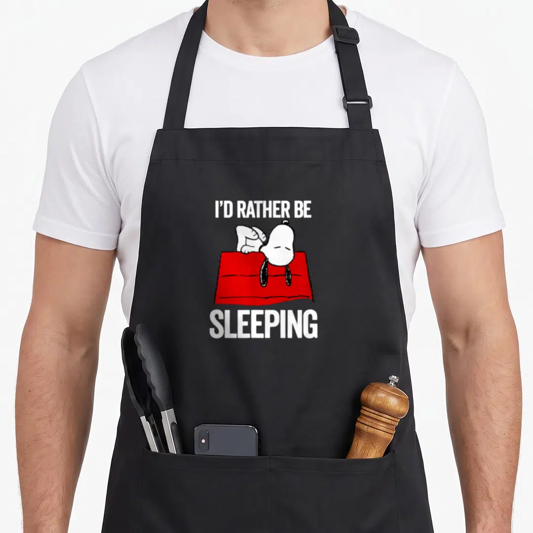 Sleeping peanut apron - Cartoon dog lying on red doghouse with white text I'D RATHER BE SLEEPING — shown on a black cotton canvas apron against a white background