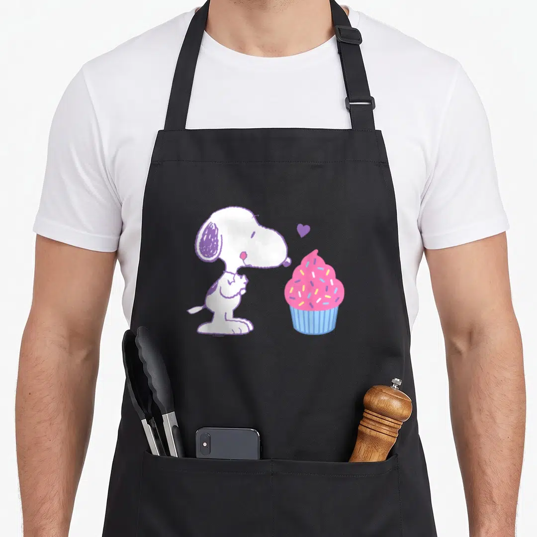 Valentine cupcake apron - Snoopy beside pink cupcake with purple heart and blue wrapper — shown on a black cotton canvas apron against a white background